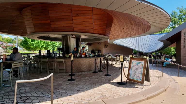 Curved wood-clad bar pavilion with a menu board, rope stanchions and patio tables on circular brick paving