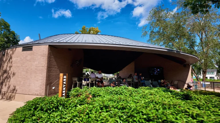 Covered patio pavilion with a sloped metal roof and a Barkyard sign, people seated at tables behind a hedge