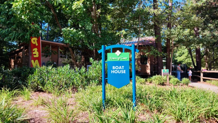 Blue Town of Cary Boat House sign in a planted bed in front of the wooden boathouse