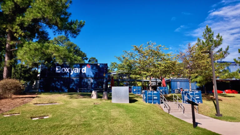 Blue shipping containers stacked into a food hall with a Boxyard RTP sign, trees and lawn in front
