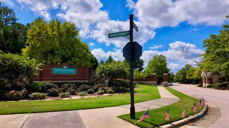 Brick entrance wall with a teal Carolina Preserve at Amberly sign, a Yates Store Rd street sign and small flags along the curb