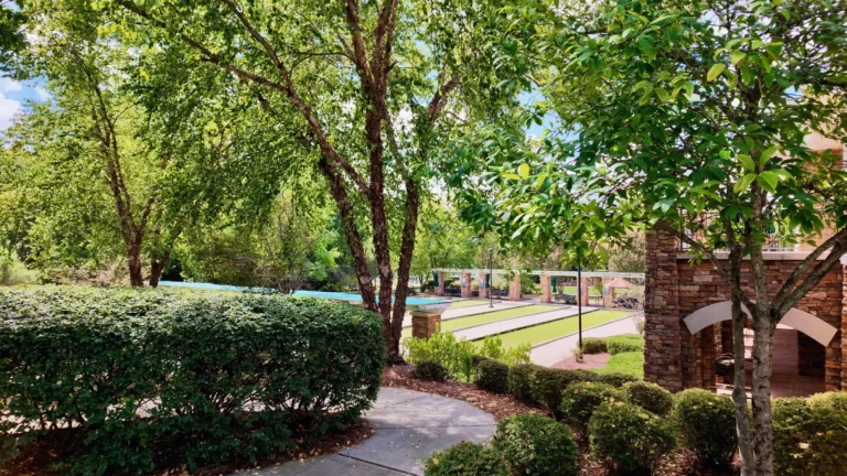 Landscaped walking path past trimmed shrubs and a brick pillar, with courts and shade structures in the background