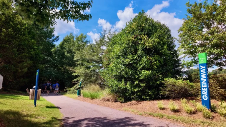 Paved greenway trail with walkers in the distance, beside a blue and green Town of Cary Greenway sign reading Cary Park Lake Trail