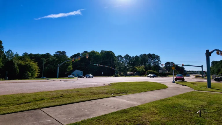 Wide signalized intersection with multiple lanes, a sidewalk and grass median, tree line behind