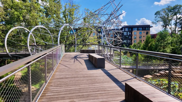 Elevated wooden skywalk with benches, passing under a sculpture of steel hoops, with an apartment building beyond