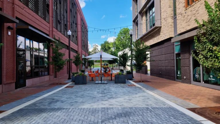 Brick-paved pedestrian passage between buildings, with string lights overhead and orange chairs under umbrellas