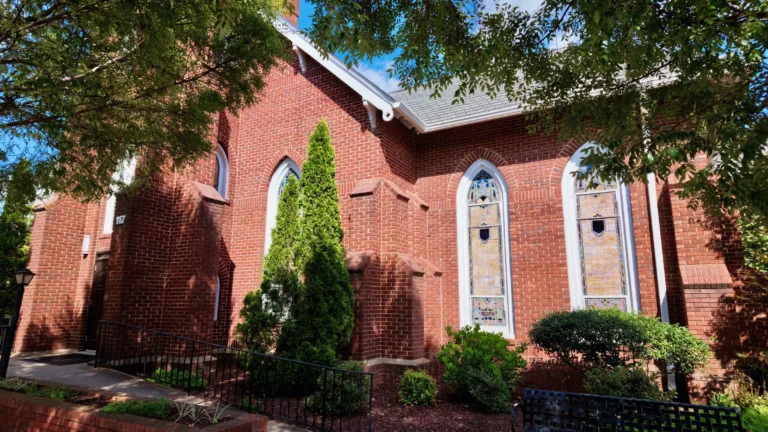 Red brick church with tall arched stained-glass windows and a narrow evergreen by the wall