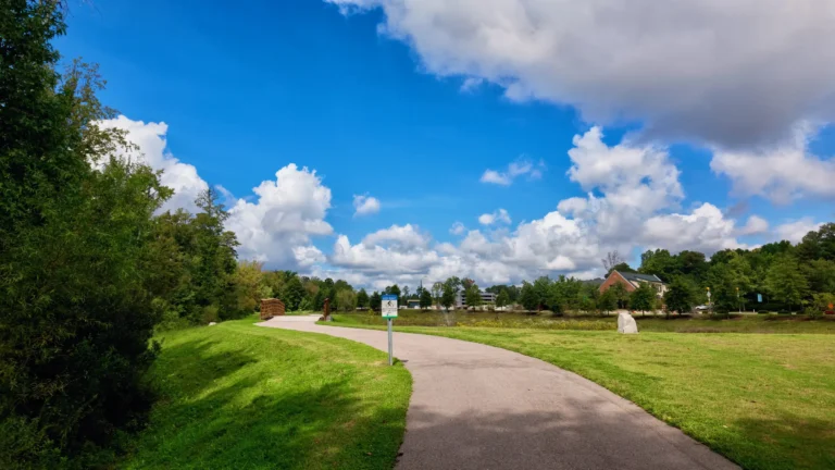 Paved greenway path curving through mowed grass and trees under a blue sky with white clouds