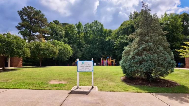 Town of Morrisville park sign reading Park Hours Dawn to Dusk on a lawn, with a fenced playground behind it