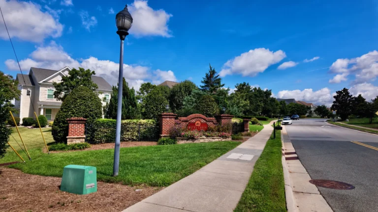 Brick entrance wall with a red Kitts Creek sign, flowering plants and a sidewalk beside the road