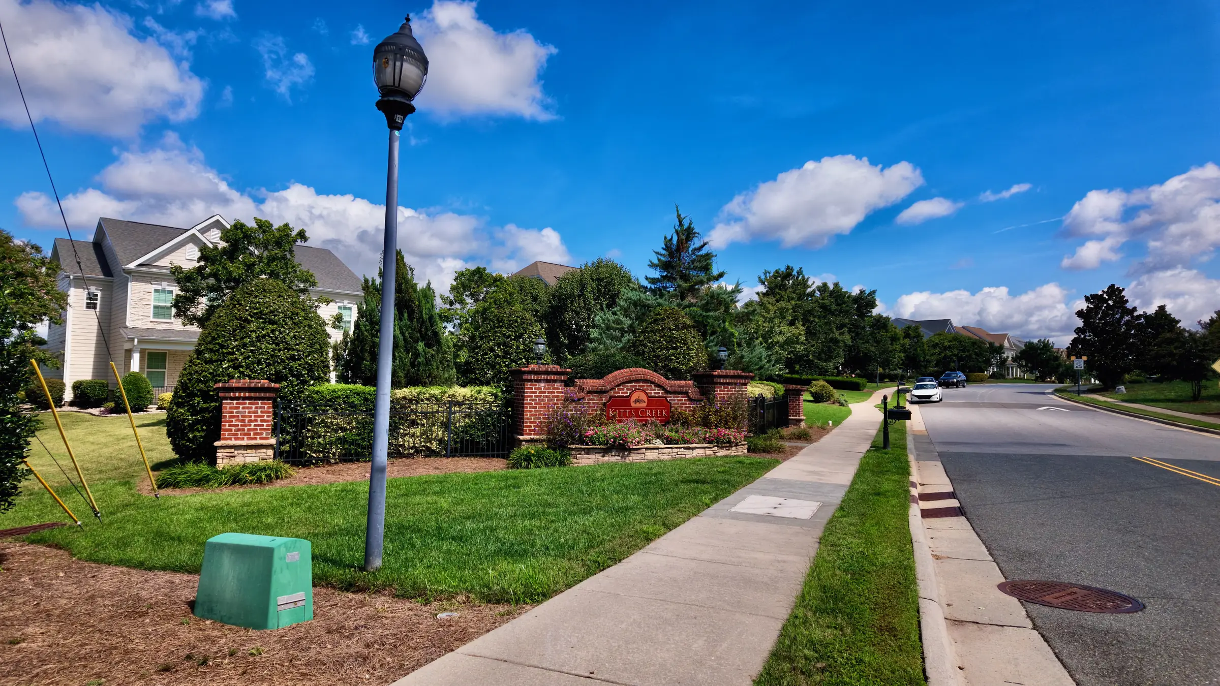 Brick entrance wall with a red Kitts Creek sign, flowering plants and a sidewalk beside the road