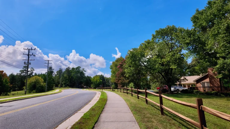 Curving two-lane neighborhood road with a sidewalk and split-rail fence, a brick ranch house on the right