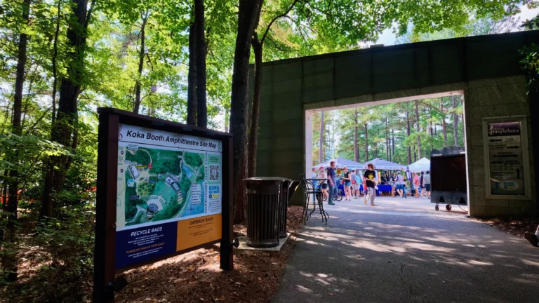 Koka Booth Amphitheatre site map sign beside an open gateway, with a crowd and vendor tents beyond