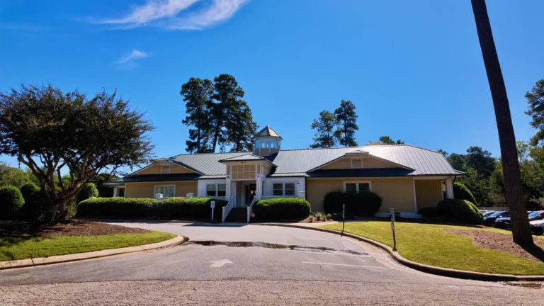 Single-story clubhouse with a green metal roof and a cupola, a curved drive and lawn in front