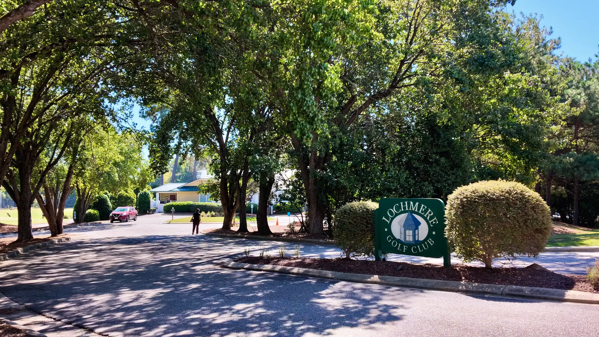 Green Lochmere Golf Club sign between two rounded shrubs, with the clubhouse drive beyond