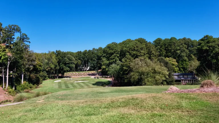 Striped fairway leading to a green with bunkers, tall trees and a house at the edge