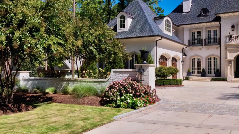Cream brick French-country house with arched doors and a paver motor court behind a garden wall
