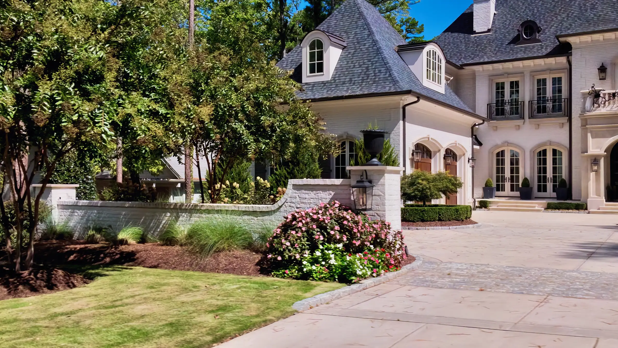 Cream brick French-country house with arched doors and a paver motor court behind a garden wall