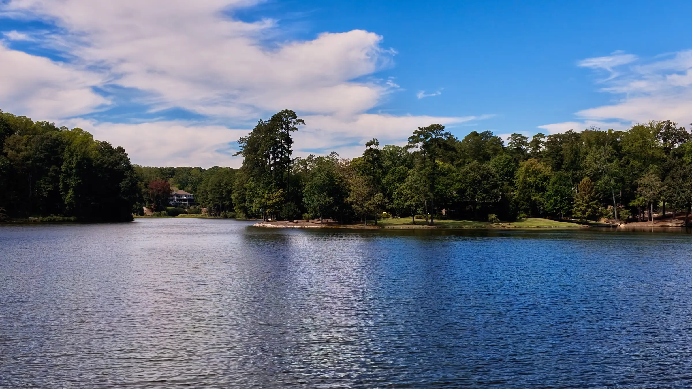 Wide lake under a partly cloudy sky with a wooded point and a home tucked in the trees