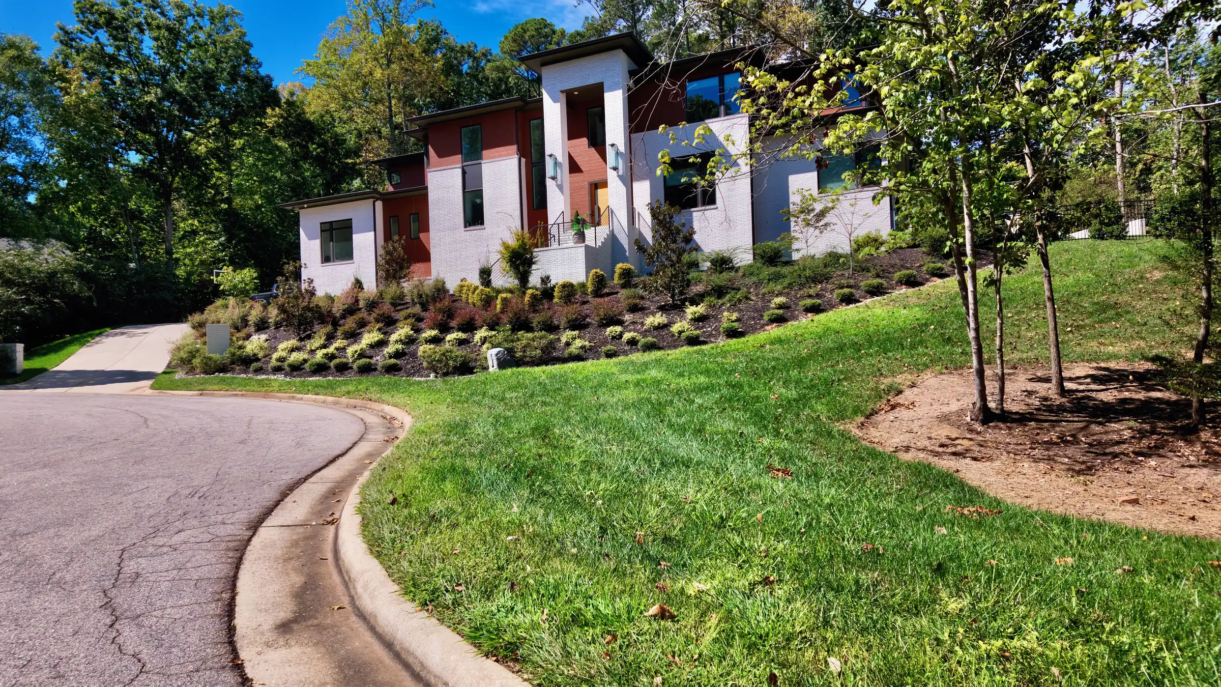 Modern two-story house with white brick and dark red panels above a landscaped slope and curving street