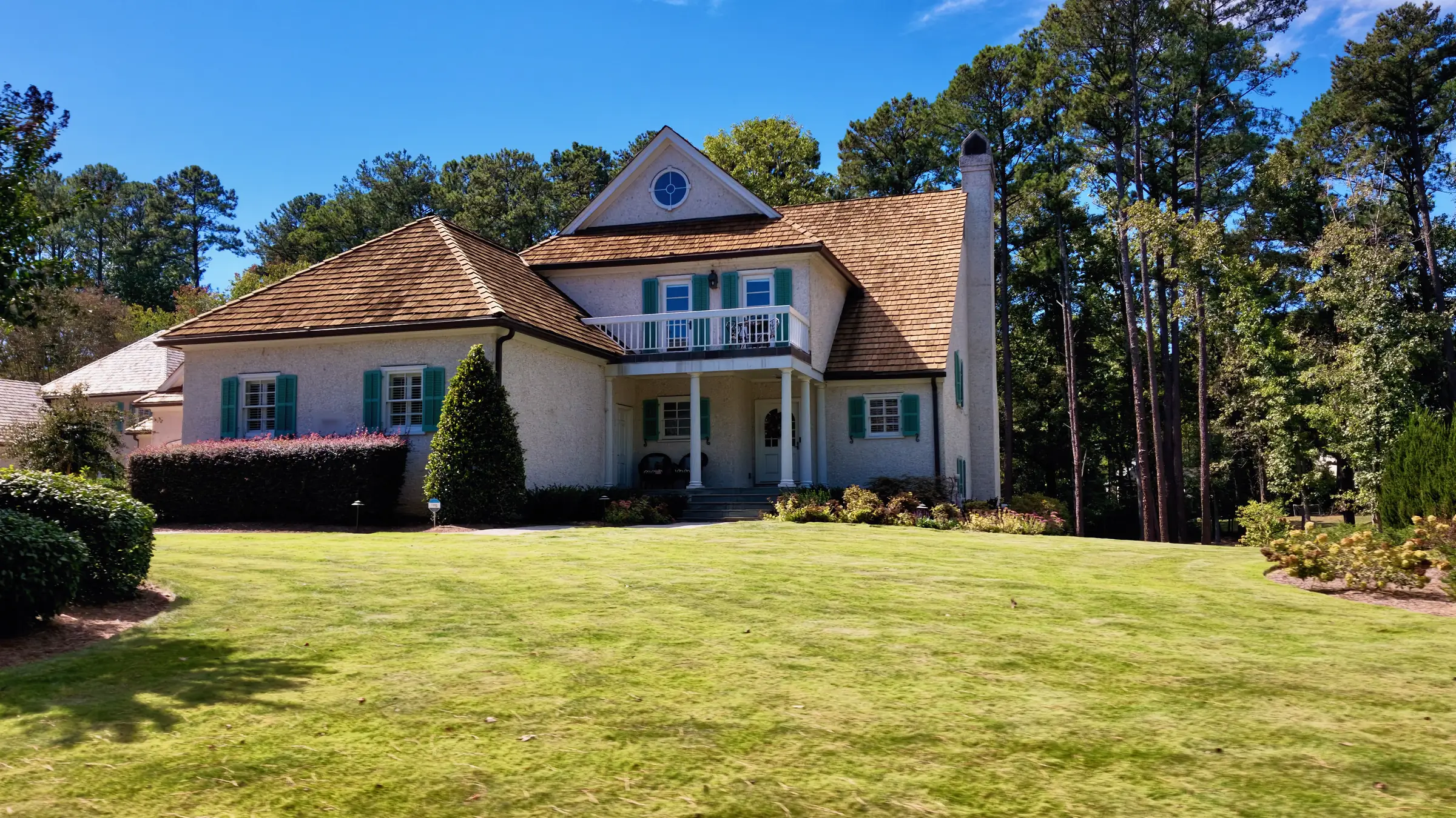 White stucco house with teal shutters, a second-floor balcony and a cedar shake roof above a wide lawn
