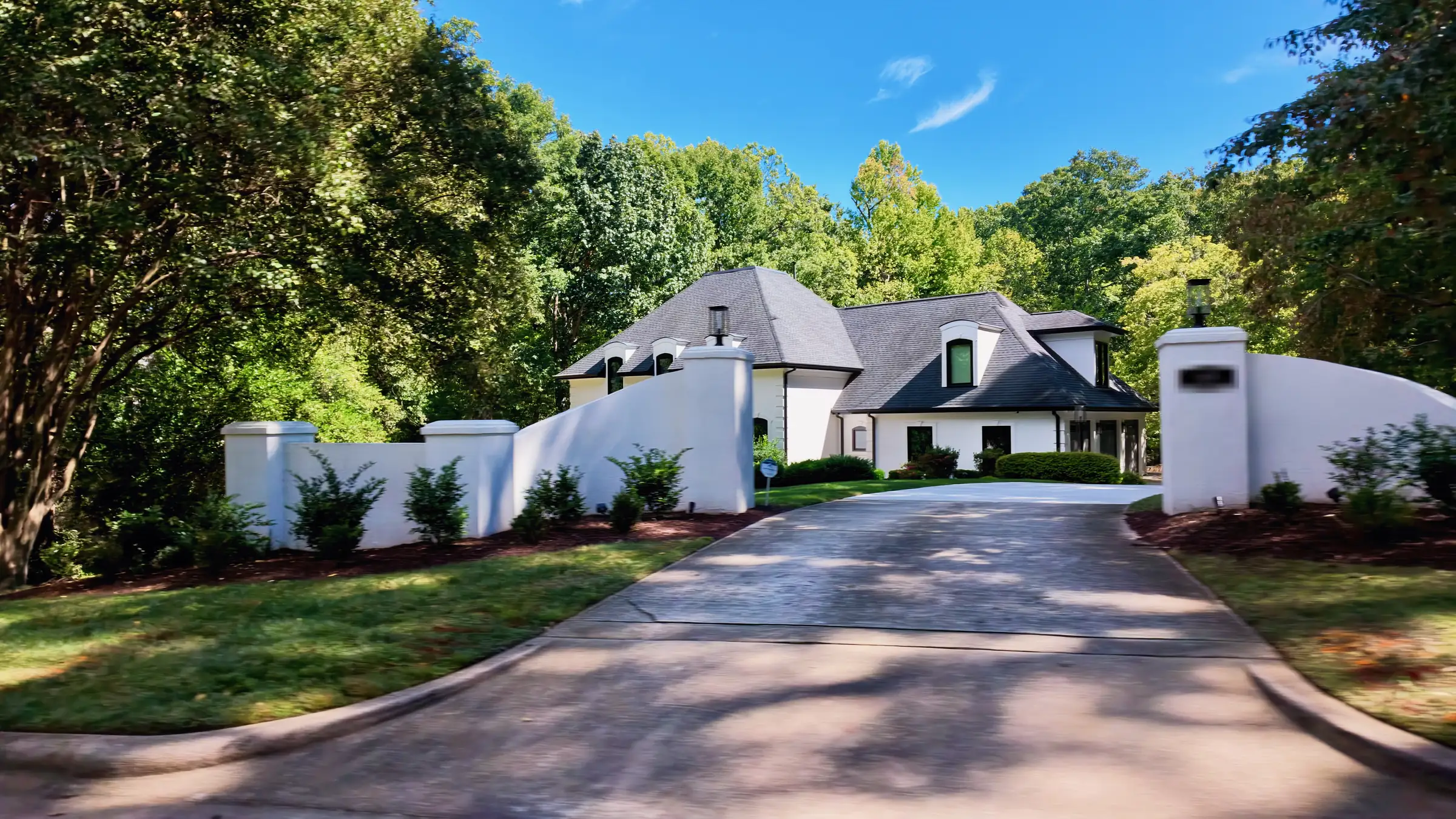 White stucco house with a steep slate-gray roof behind curved walls and pillars at a wide driveway