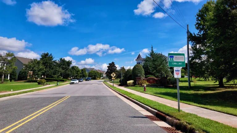 Green Morrisville Town Limit sign beside a two-lane road, with the Kitts Creek entrance wall and a turreted house behind it