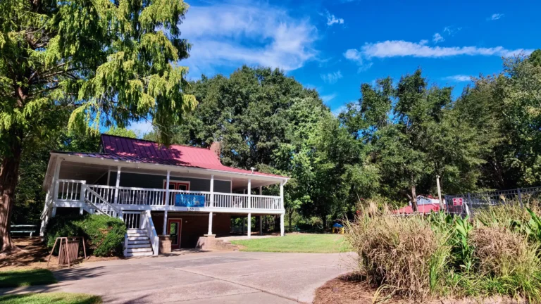 Clubhouse with a red metal roof and white wraparound porch with an Oxxford Hunt sign, big trees behind