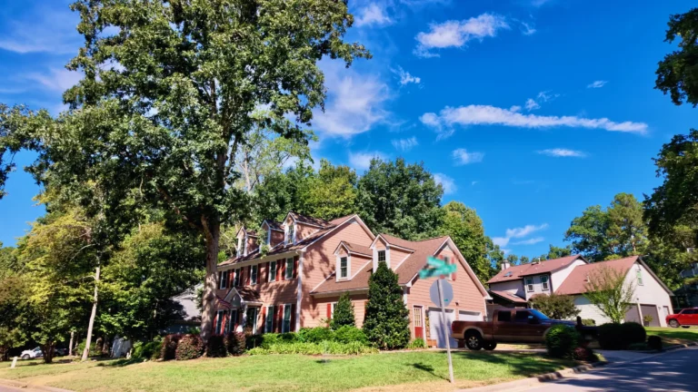 Two-story salmon-colored home with dormers and a two-car garage on a corner lot under tall trees
