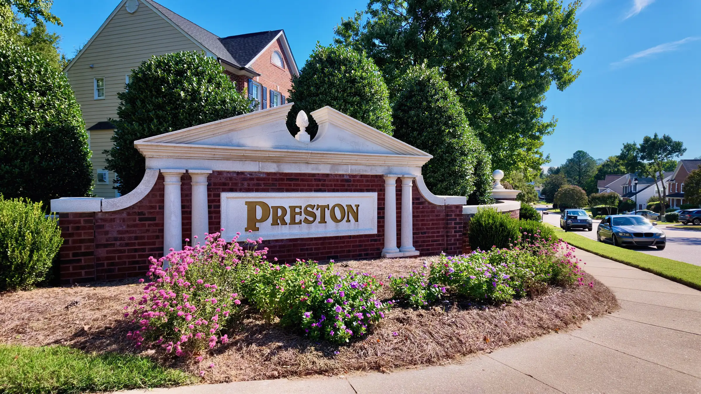 Brick entrance wall with white columns, a pediment and gold Preston lettering behind pink and purple flowers
