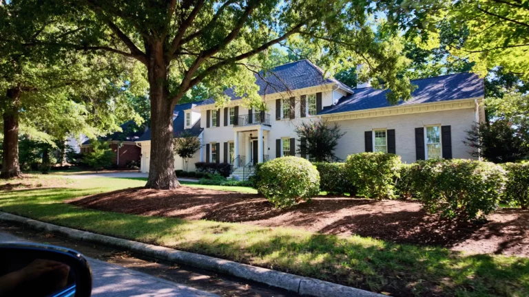 Two-story painted white brick house with black shutters and a small balcony behind a large oak and trimmed shrubs