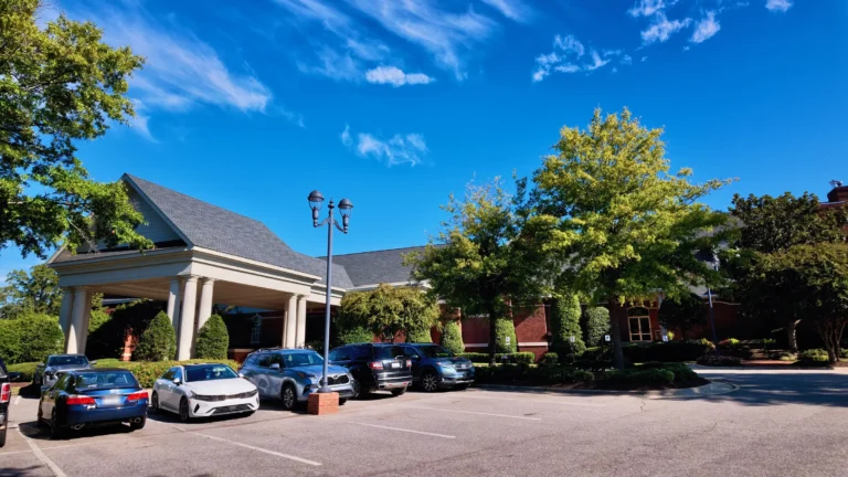 Brick clubhouse with a white-columned covered entrance, a two-headed lamppost and cars in the lot