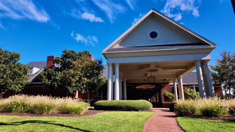 White-columned porte-cochere with a round window in the gable, a brick walk and ornamental grasses