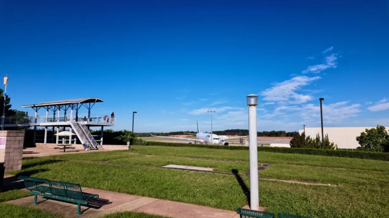 Open lawn with benches and the viewing deck at left, a jet taxiing on the far runway