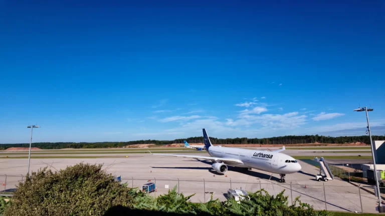A Lufthansa wide-body jet parked at an RDU gate, seen over shrubs and a fence from the observation park
