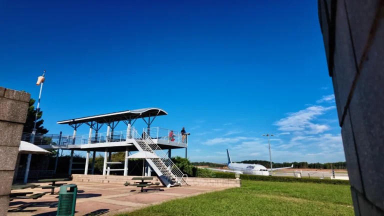 Two-level steel viewing deck with a curved roof on a lawn, a jet on the runway behind it
