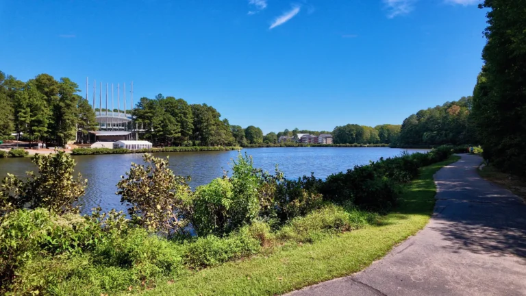 Paved greenway curving along a lake past shrubs, the amphitheater canopy in the distance