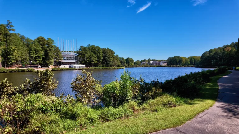 Lake with a paved path along the shore and the amphitheater's white canopy masts on the far bank