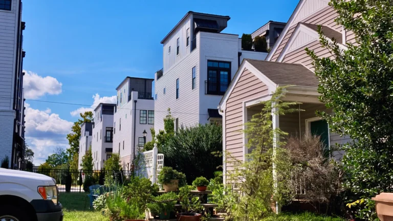 Row of tall white modern townhomes next to an older cottage with a porch and potted plants