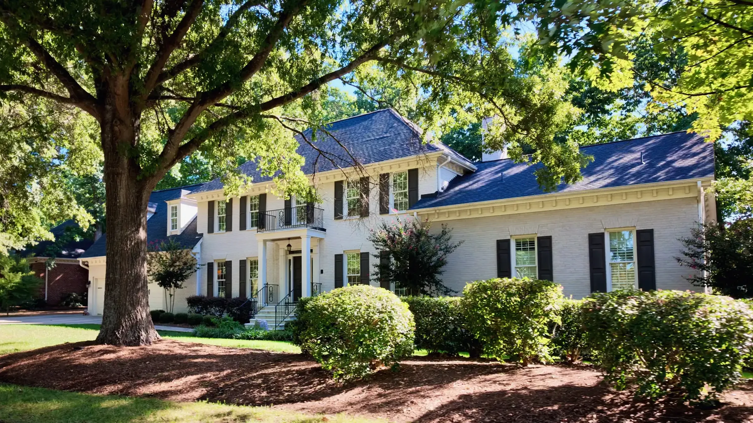Two-story painted white brick house with black shutters and a small balcony behind a large oak and trimmed shrubs